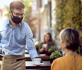 Camarero con mascarilla y guantes llevando dos cafés a clientas